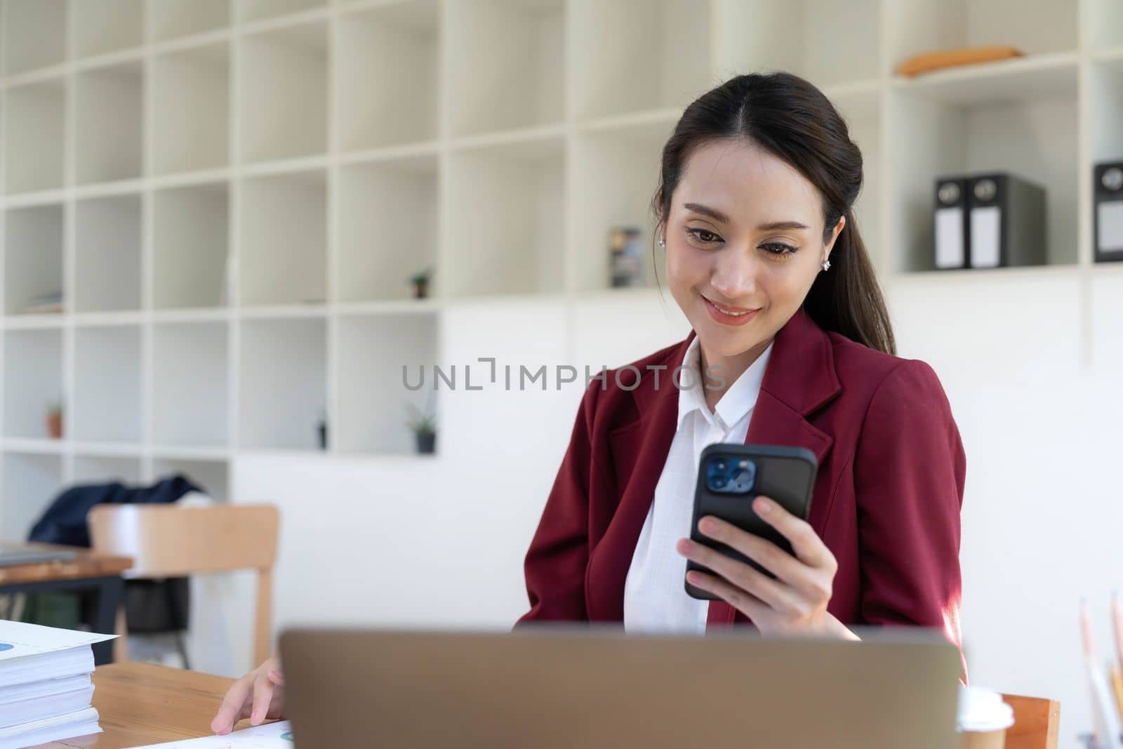 Smiling beautiful Asian businesswoman analyzing chart and graph showing changes on the market and holding smartphone at office..