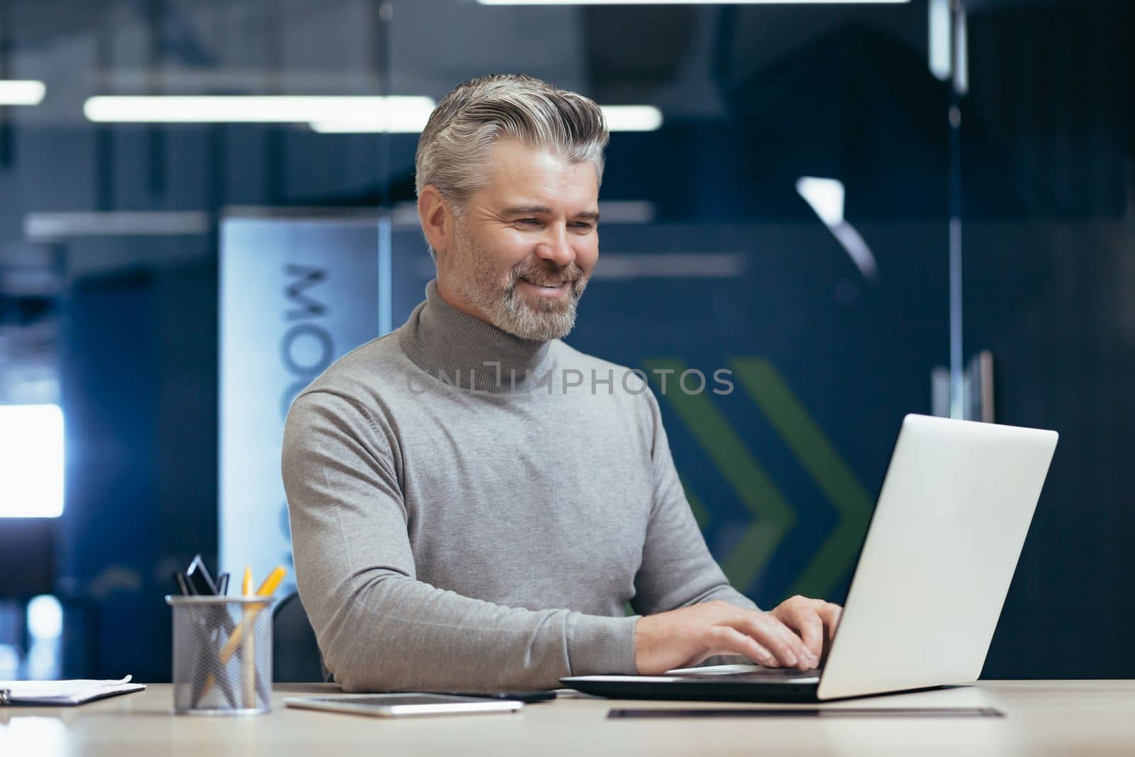 Mature successful boss working inside modern office, senior gray haired businessman at work working with laptop smiling and happy investor happy with achievement.