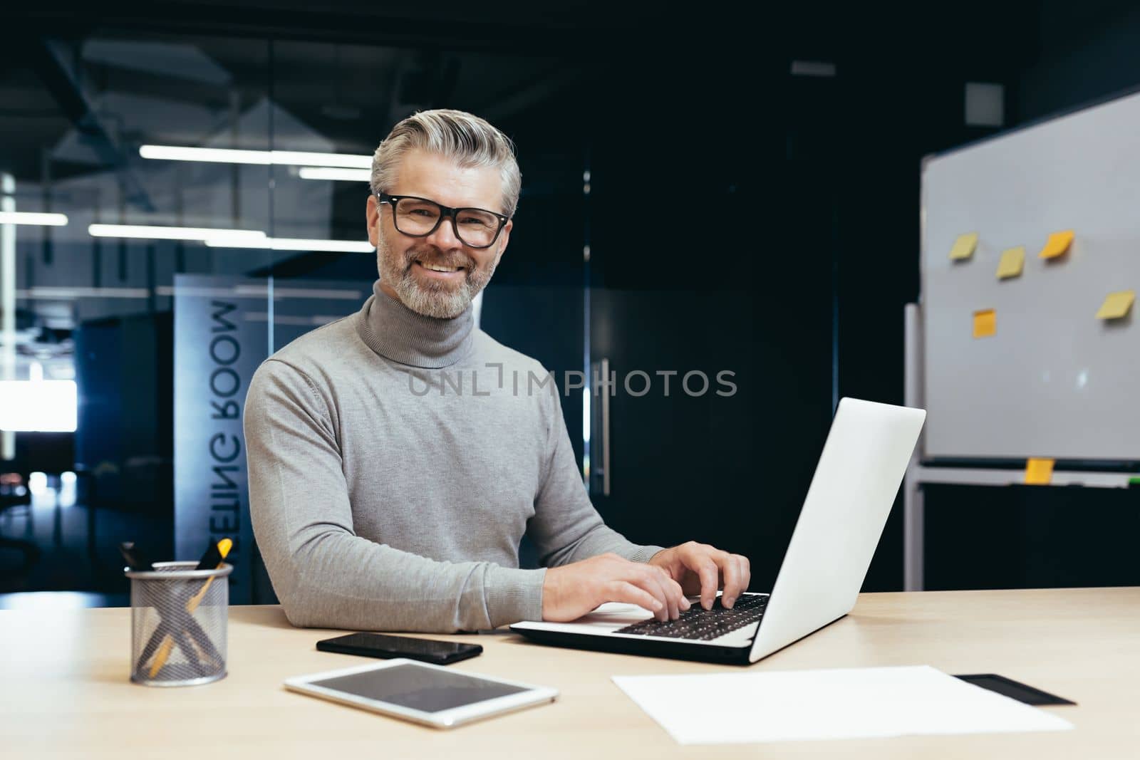 Portrait of successful senior businessman, gray haired man in glasses smiling and looking at camera, mature investor boss working inside modern office building using laptop at work.