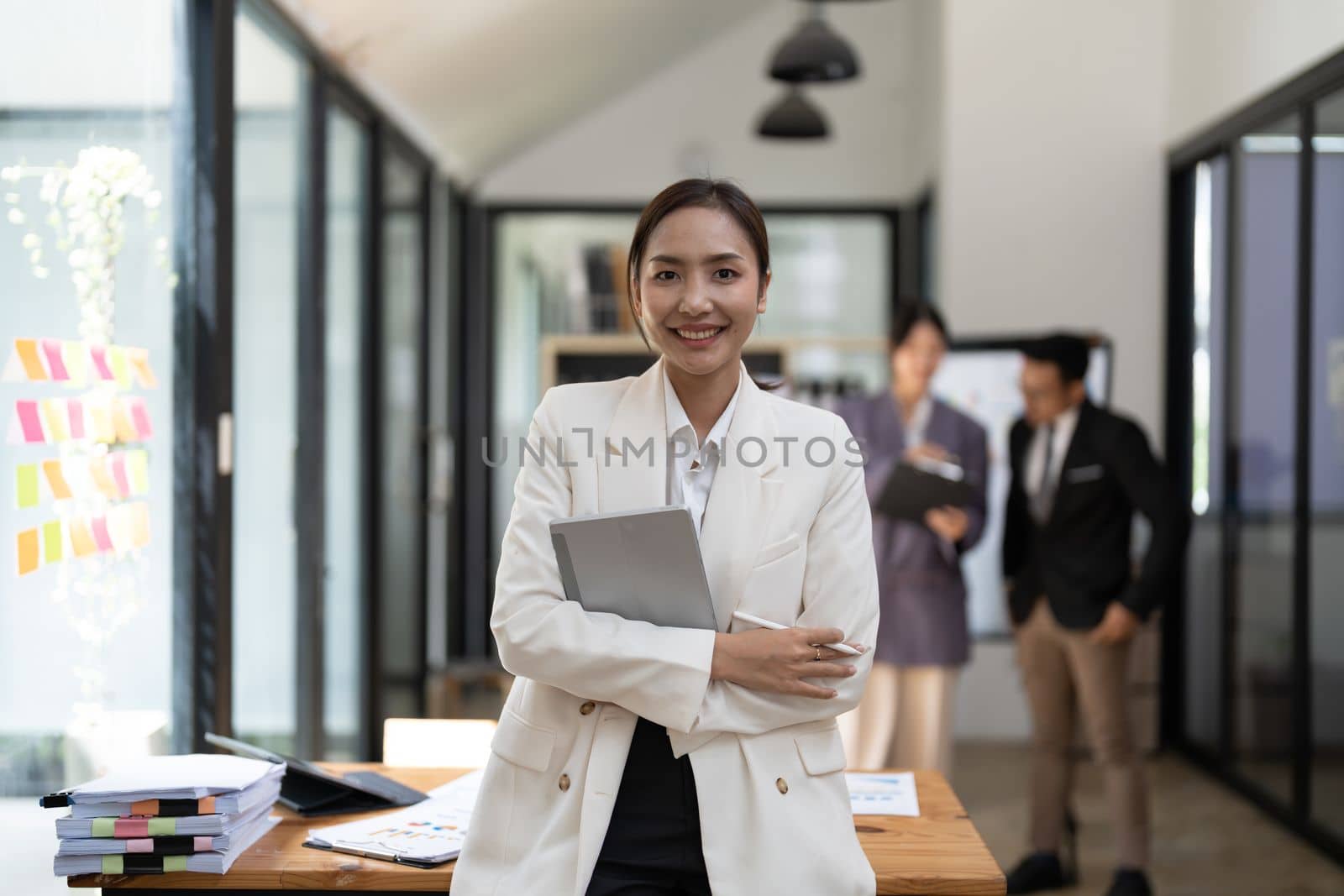 Portrait of young attractive asian female office worker in formal business suits smiling at camera in office with blurry colleagues working in office as background. by nateemee