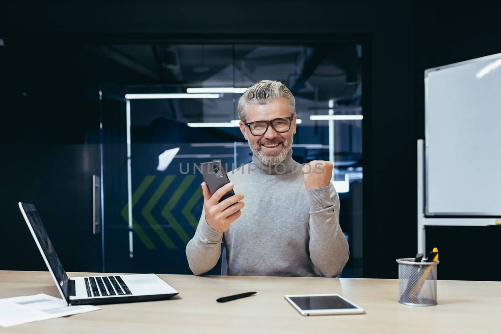 Businessman celebrating victory and success, mature gray haired man inside office got good news result using smartphone, man at work with laptop.