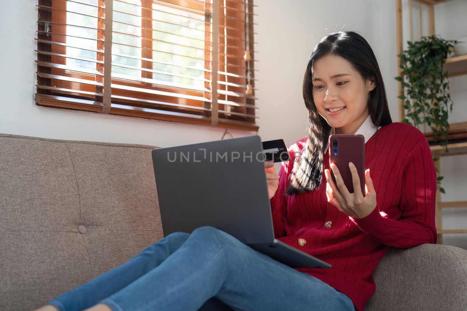 Beautiful young woman holding credit card while using laptop computer and mobile phone sitting at the living room. Black friday online shopping concept by nateemee