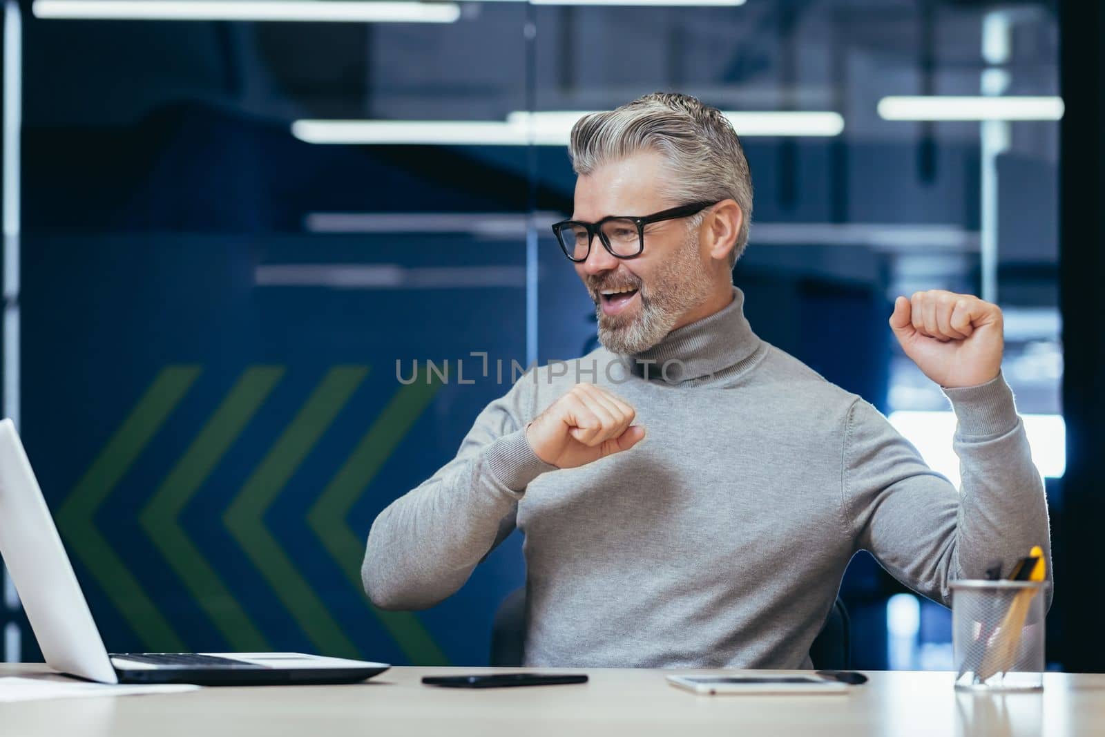 Successful gray haired boss working inside modern office, mature businessman celebrating victory looking at monitor screen and satisfied with achievement, senior man smiling and dancing sitting.