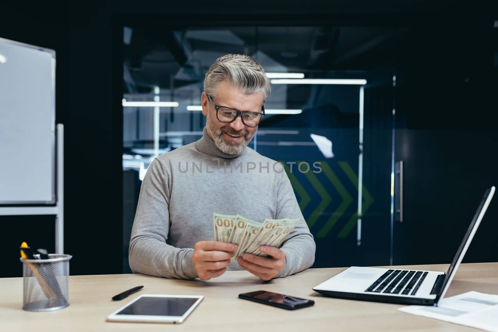 Mature successful financier investor counts money dollars cash sitting inside office at table, senior businessman happy with result achievement and income uses laptop at work.