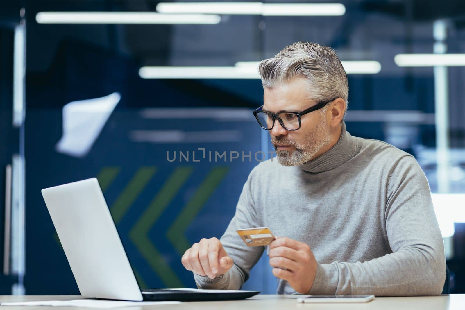 Senior gray haired businessman in office frustrated and sad trying to make bank transaction and purchase in online internet store, man holding bank credit card and using laptop.