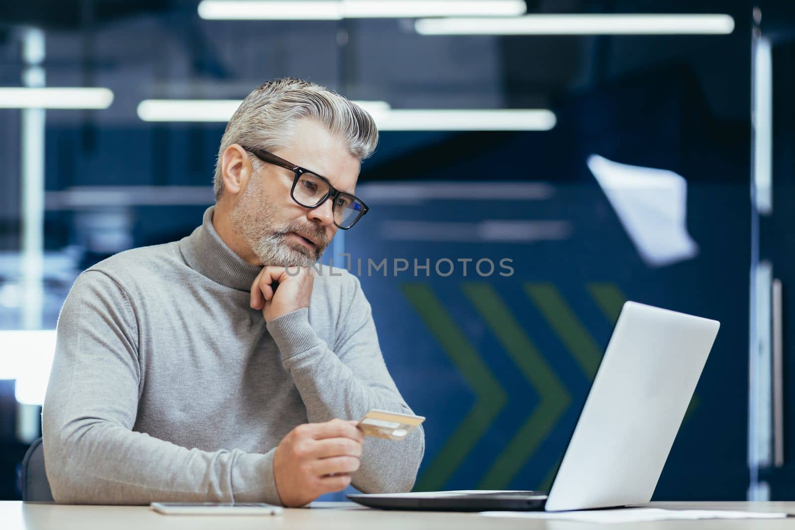 Senior gray haired businessman in office frustrated and sad trying to make bank transaction and purchase in online internet store, man holding bank credit card and using laptop.