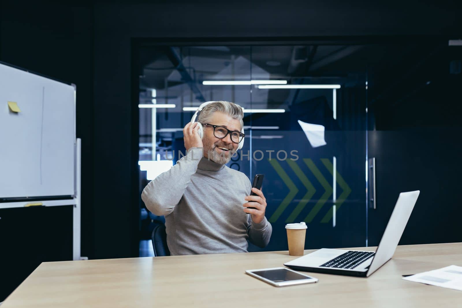 Senior gray haired businessman boss working inside modern office using laptop at work, mature man in headphones listening to music and audiobooks podcasts using app on phone.