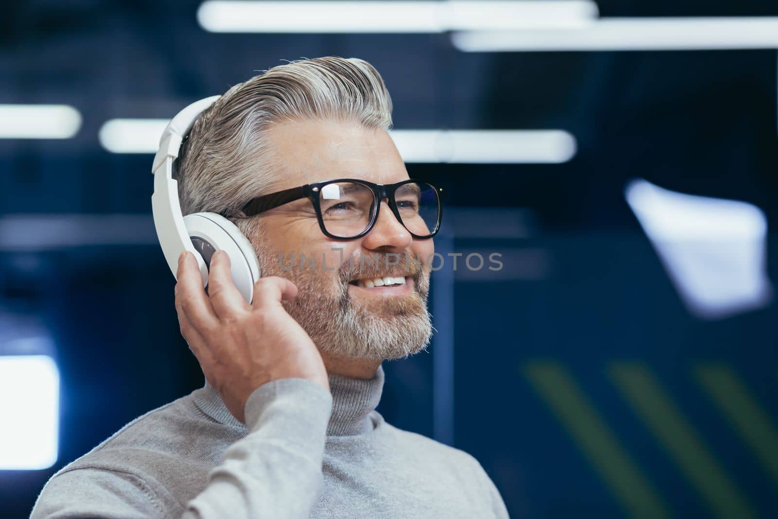 Close-up photo. Portrait of an older gray-haired man in glasses listening to music in headphones in the office, holding headphones in his hand, smiling, looking away.