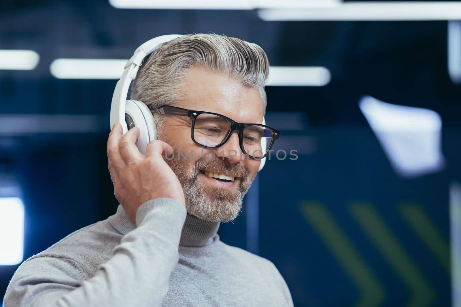 Close-up photo portrait of mature gray-haired businessman, gray-haired man in headphones listening to music audio books and podcasts online, boss at workplace inside office.