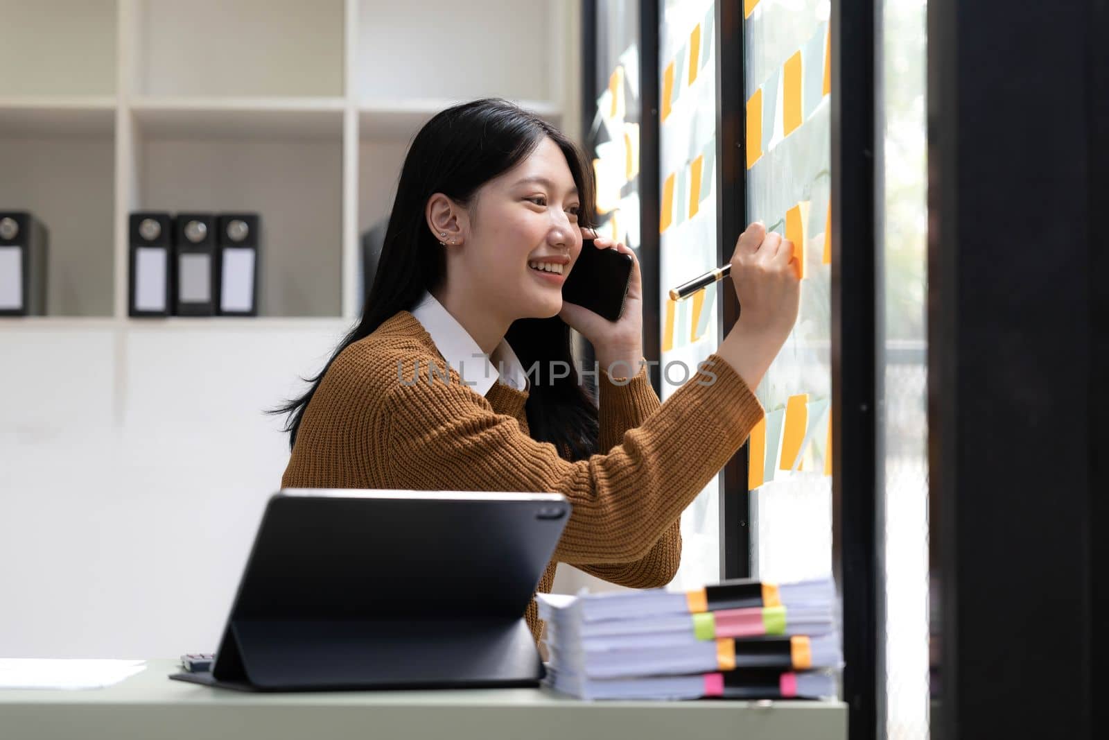 Smiling beautiful Asian businesswoman analyzing chart and graph showing changes on the market and holding smartphone at office...
