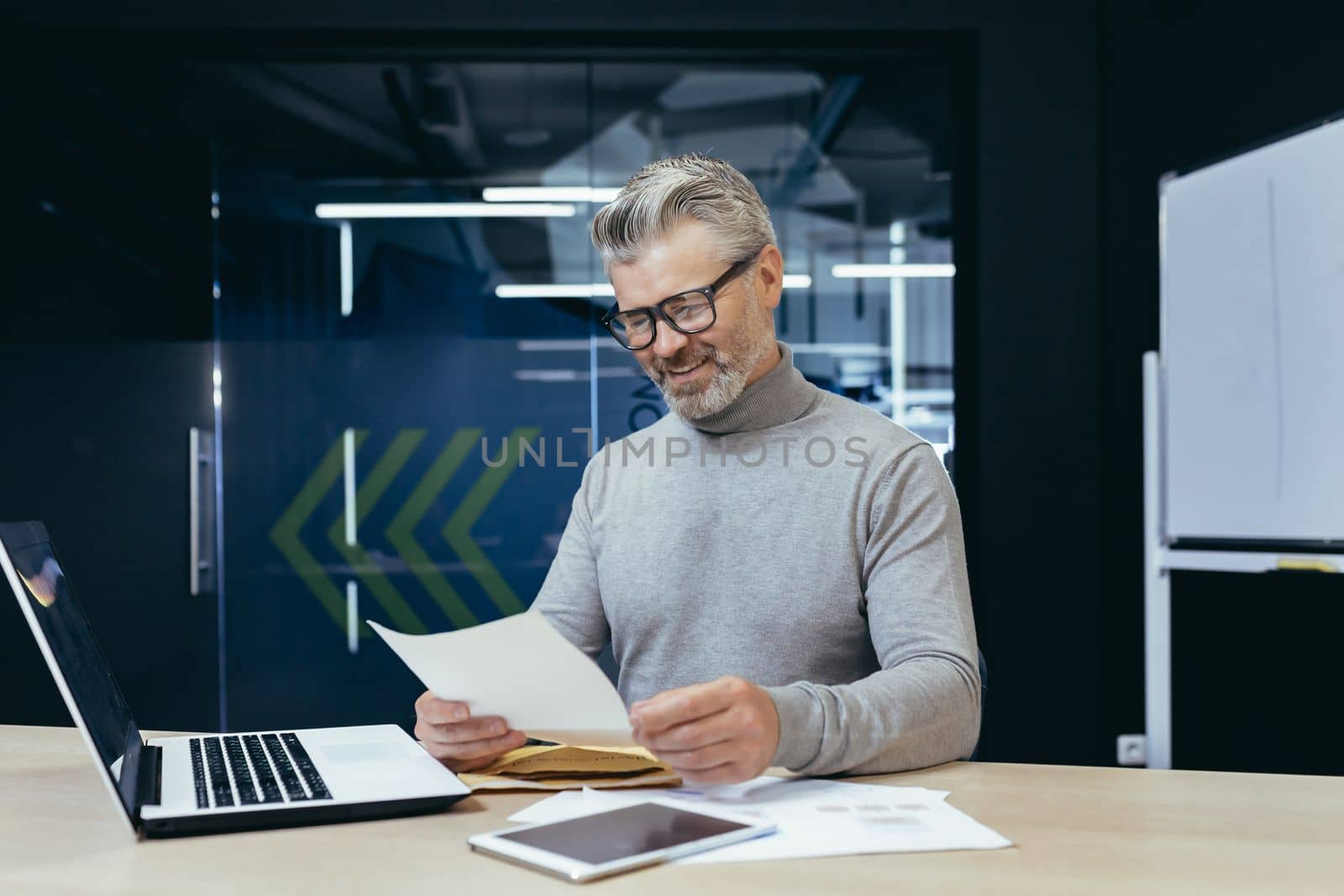 Mature successful gray haired businessman smiling and happy received letter notification, senior male boss working inside office using laptop reading good news in letter.