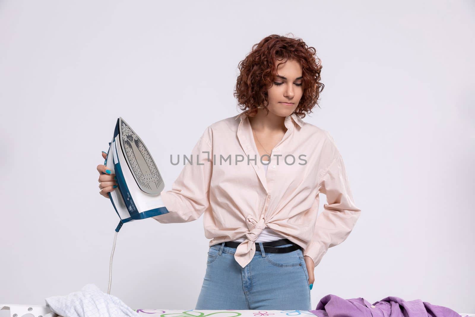 Annoyed woman with the next batch of clothes to iron. She dislikes ironing. Daily housework after washing clothes is ironing shirts.
