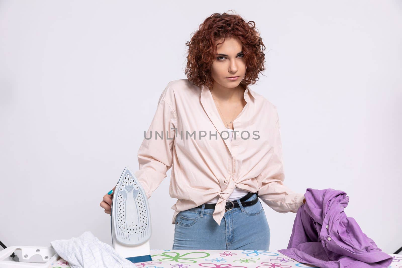 Annoyed woman with the next batch of clothes to iron. She dislikes ironing. Daily housework after washing clothes is ironing shirts.