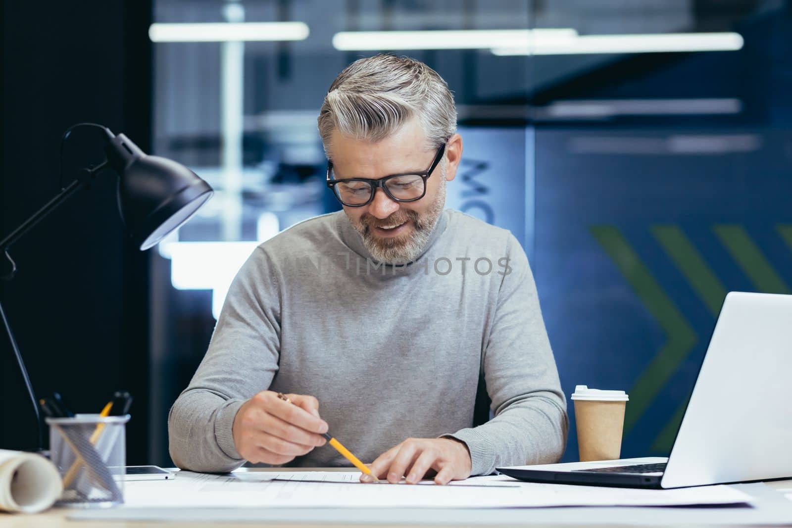 Successful senior architect at work in office, mature gray haired man drawing blueprints on layout and plan smiling and happy with work.