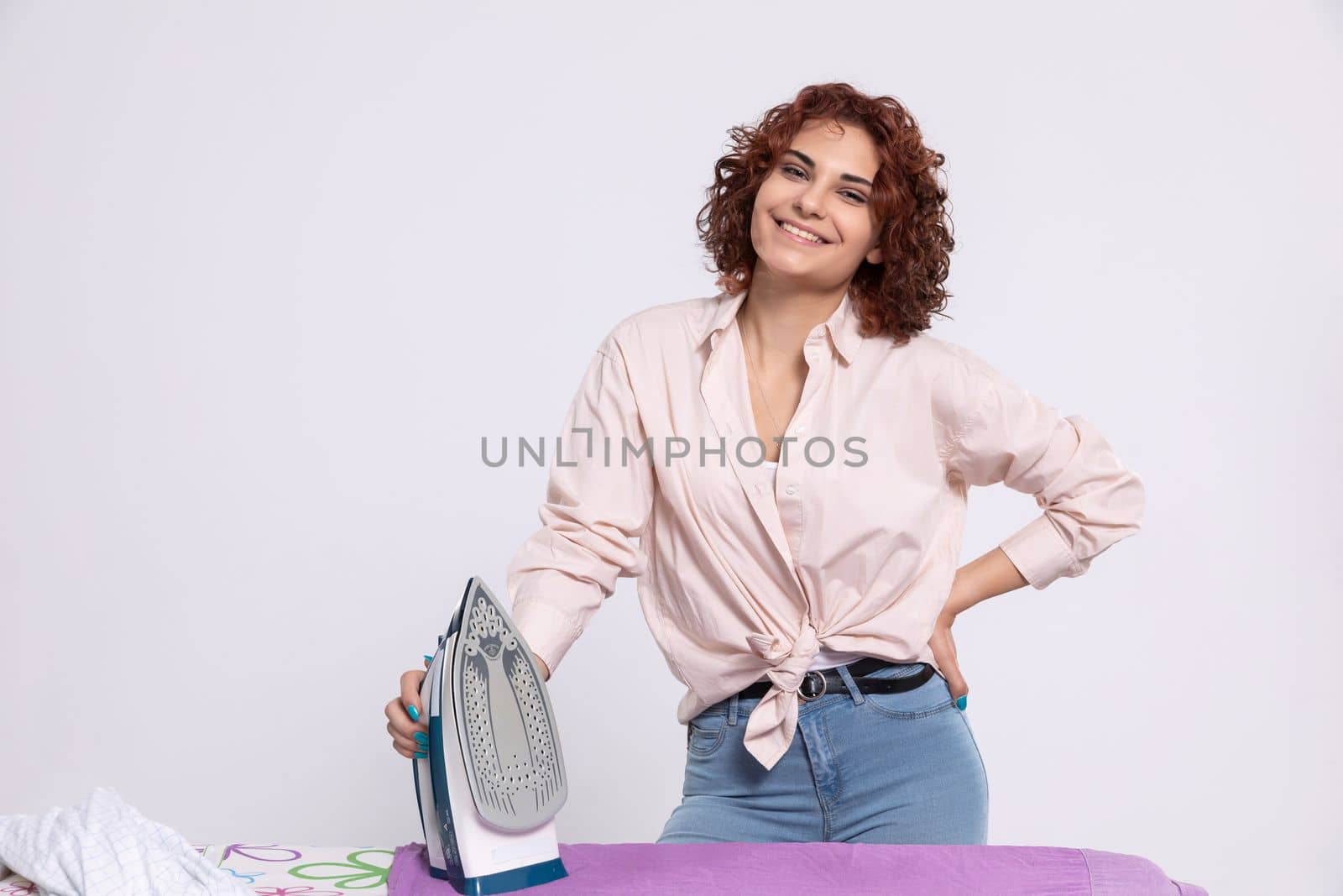 A girl with a smile on her face stands at the ironing board and in her hand holds an iron. Daily household chores.