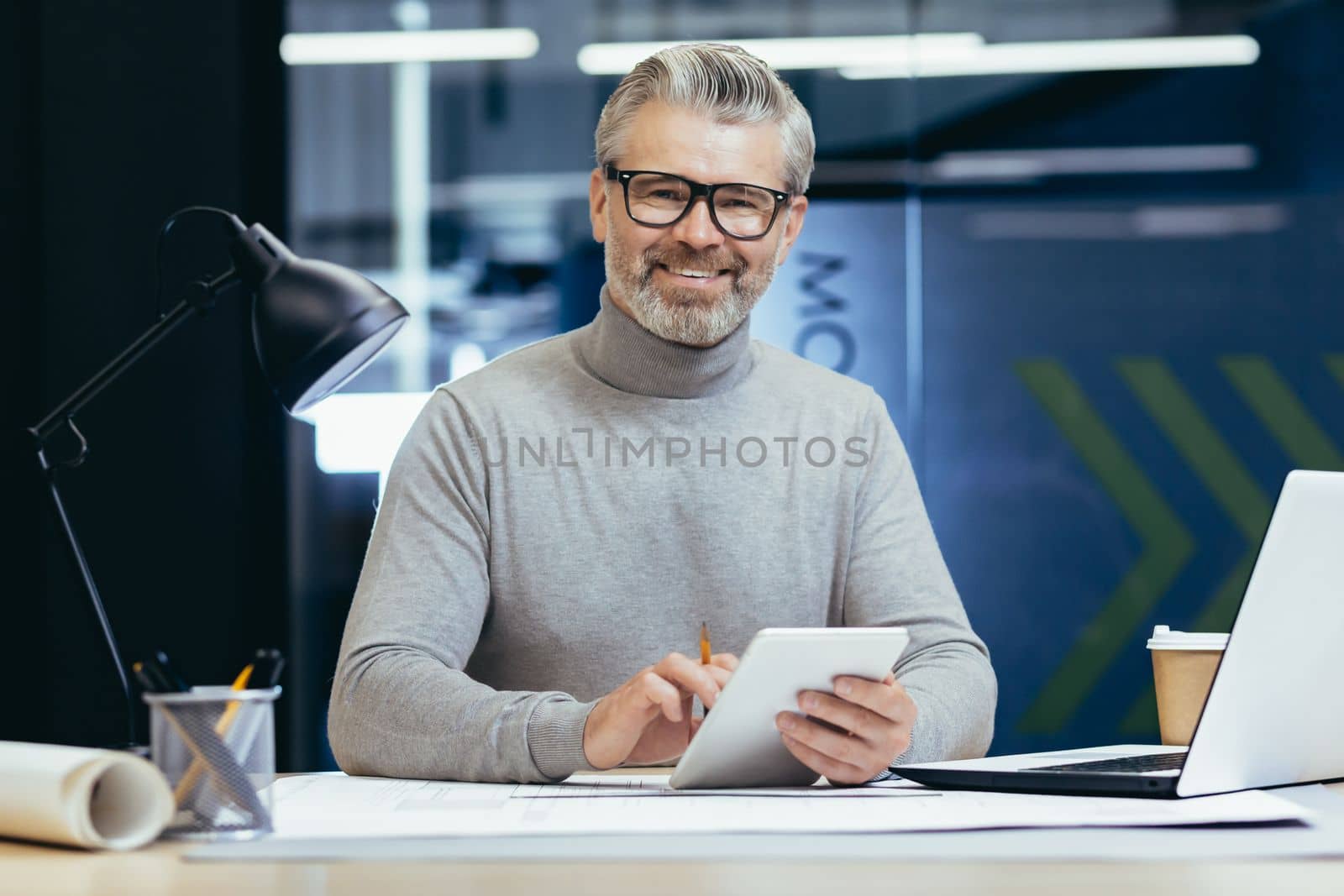 Portrait of senior architect, gray haired designer smiling and looking at camera, man mock up plan use laptop and tablet computer at work inside studio office.