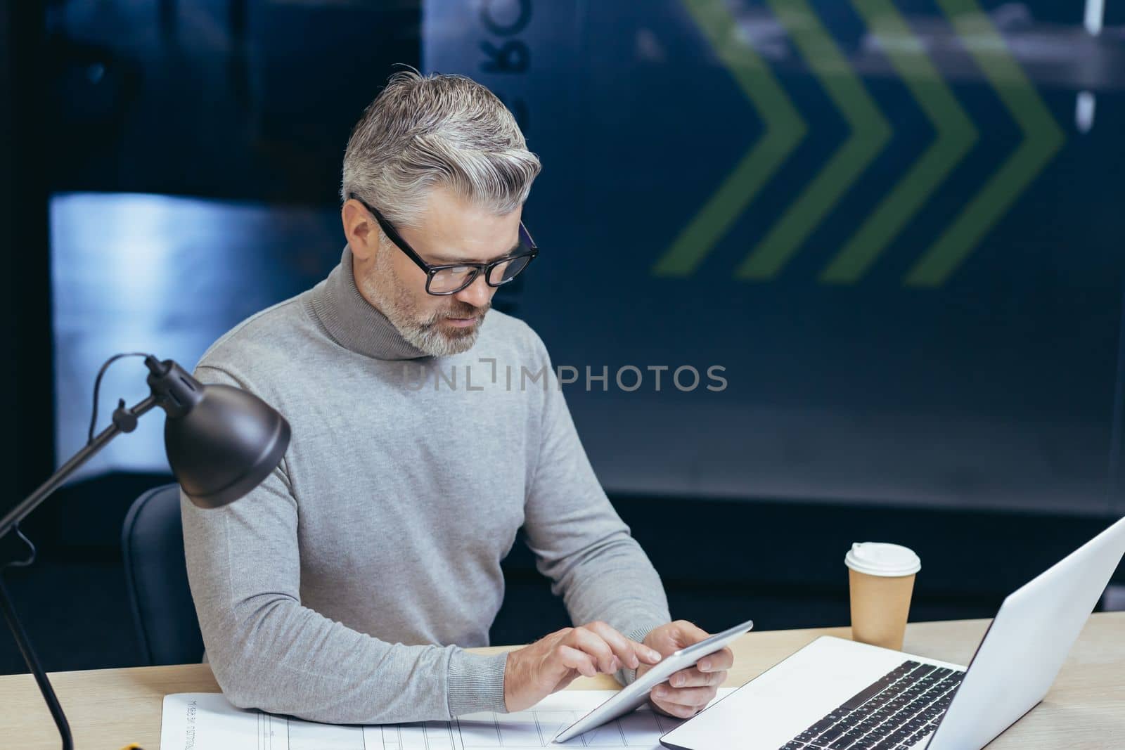 Serious thinking gray-haired businessman working inside office with laptop, senior man using tablet computer browsing internet pages reading online news.