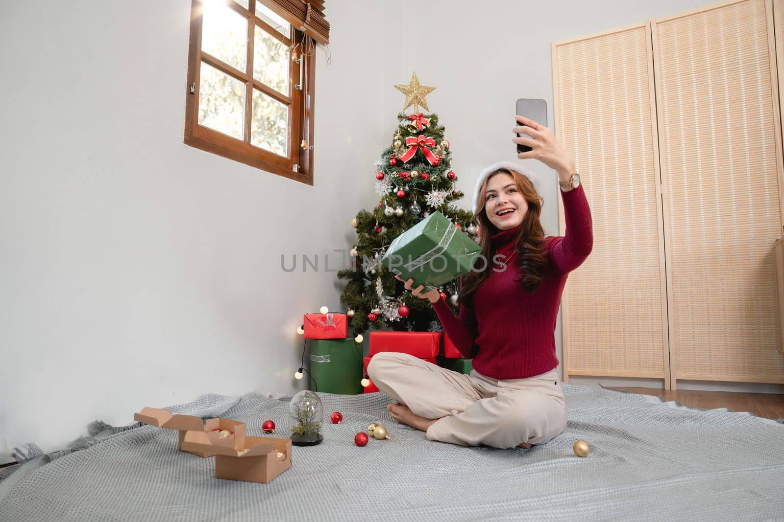 Holiday selfie at Christmas tree. Happy young girl taking selfie on smart phone at decorated Christmas tree iat home.