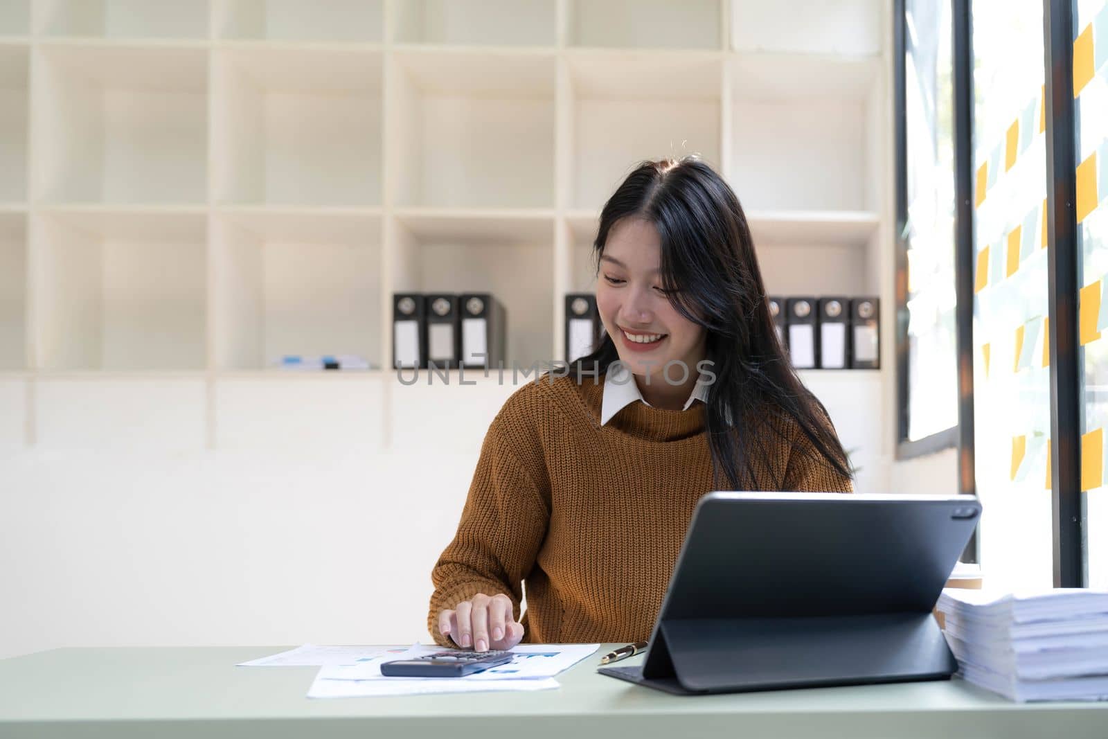 Asian Business woman using calculator and laptop for doing math finance on an office desk, tax, report, accounting, statistics, and analytical research concept..