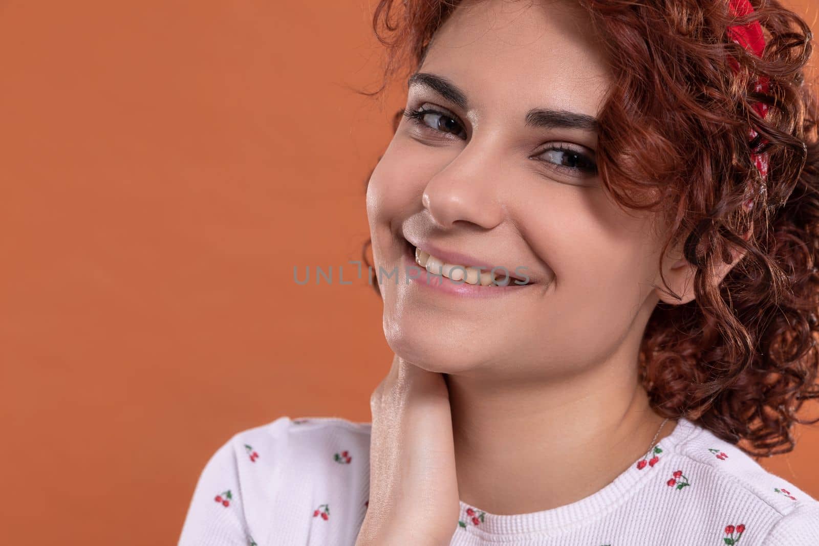 European beauty girl with short and curly hair. Uniform background in brick-red color. She is wearing a white blouse with a cherry pattern.