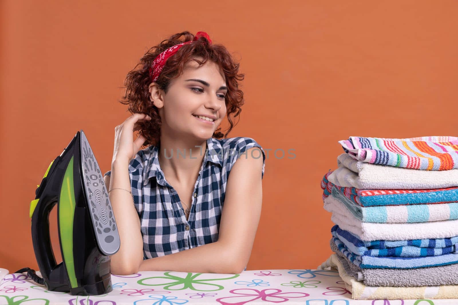 Full of joy and happiness, the girl with curly hair looks nonchalantly at the ironed towels lying on the ironing board.