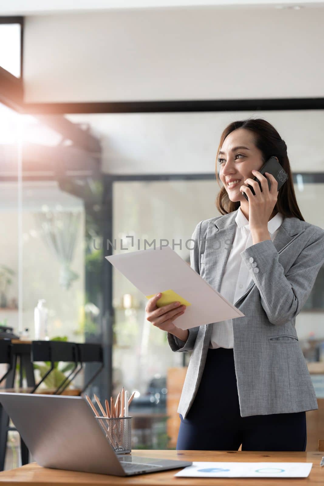 Happy young asian business woman wearing suit holding mobile phone standing in her workstation office. 