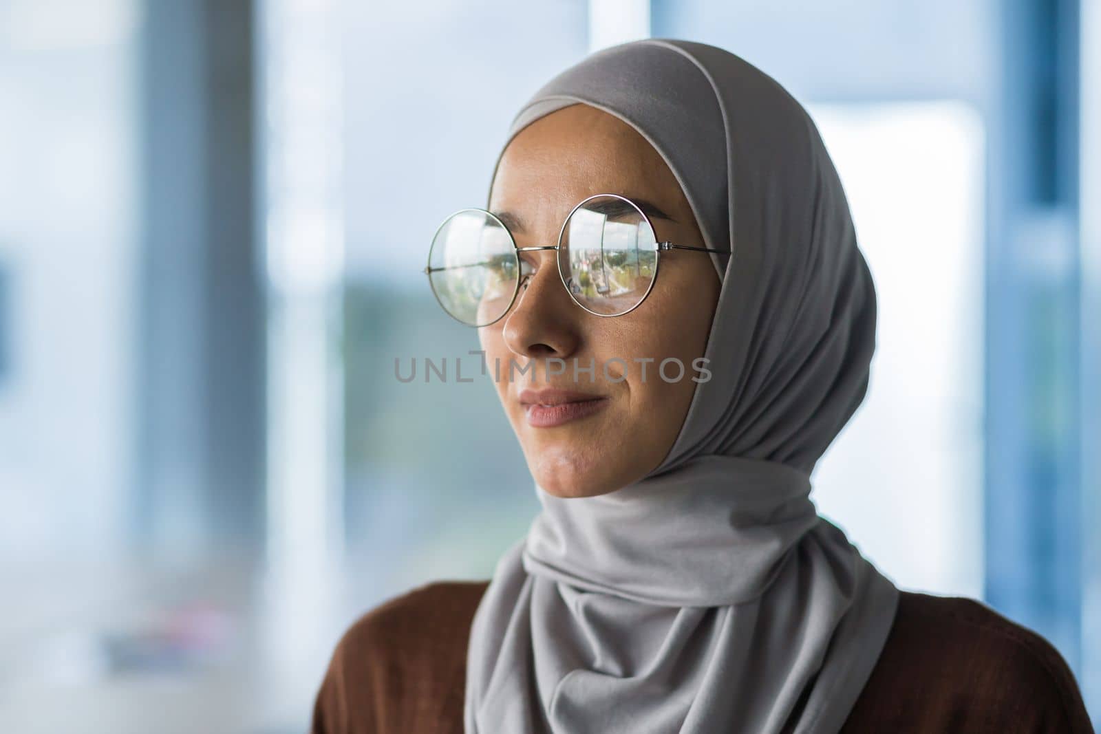 Close-up photo portrait of Muslim business woman wearing hijab and glasses, office worker looking out window thinking about decision, female boss inside office.
