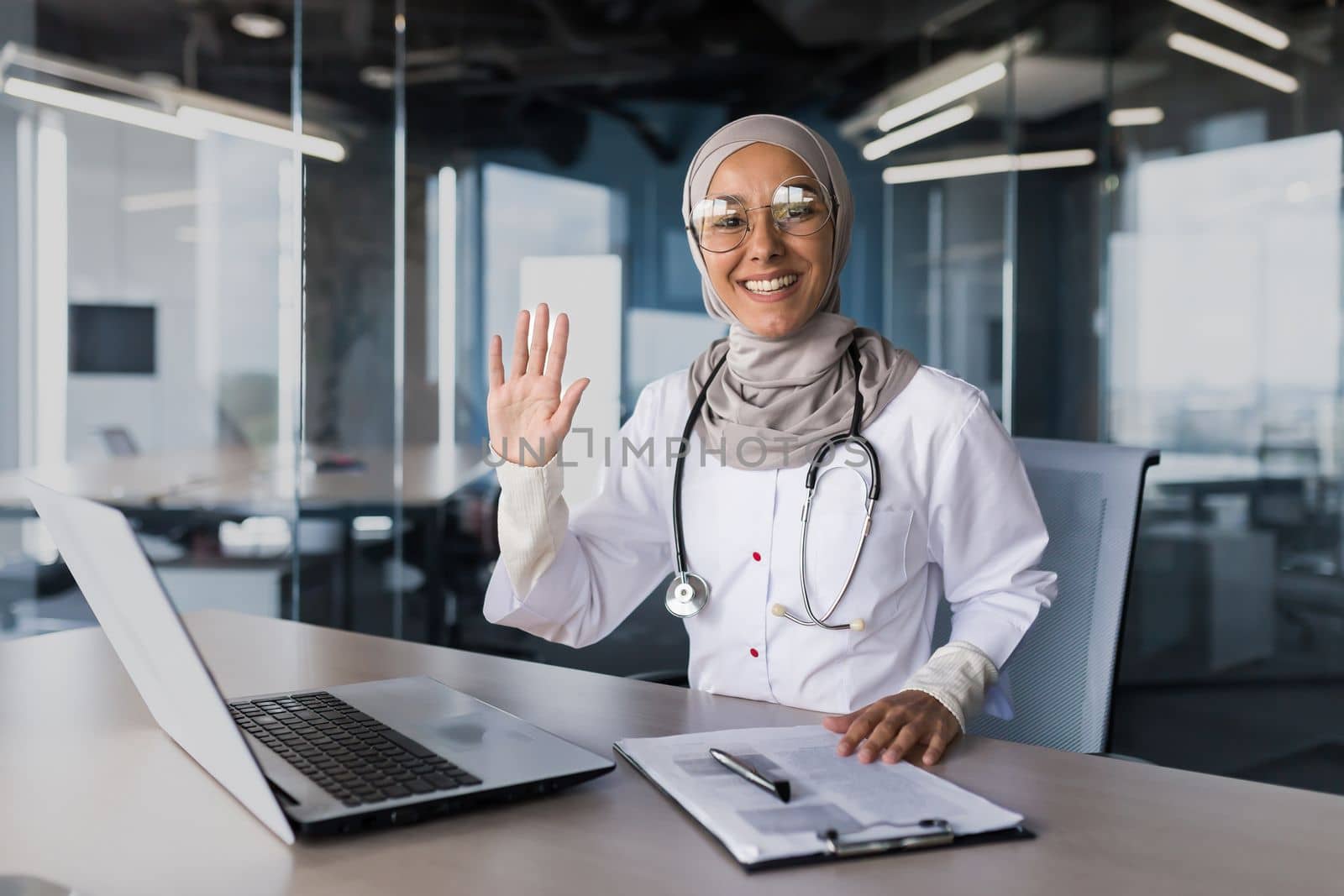 A young beautiful Arab woman, a Muslim doctor is sitting in the office at the desk in a hijab. He waves his hand at the camera, greets his patients, smiles.