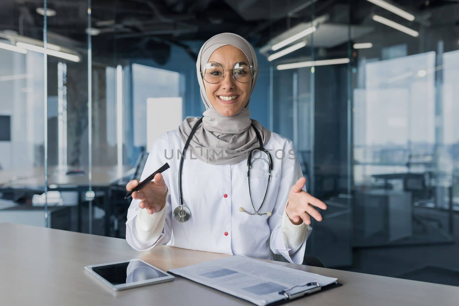 A young beautiful Arab woman, a Muslim doctor is sitting in the office at the desk in a hijab. Conducts a medical online webinar, looks into the camera, explains, smiles.