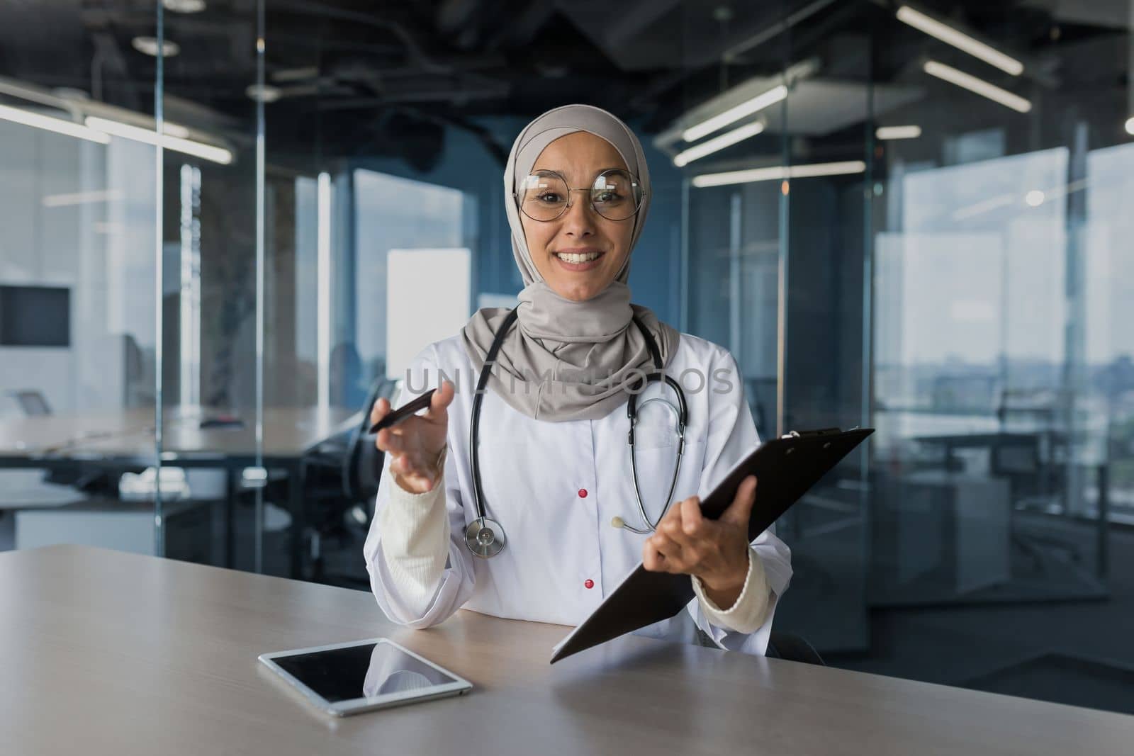 Online doctor's appointment. A young beautiful woman doctor, a Muslim woman, consults a patient remotely through a video call. He looks into the camera, explains, smiles, writes down complaints.