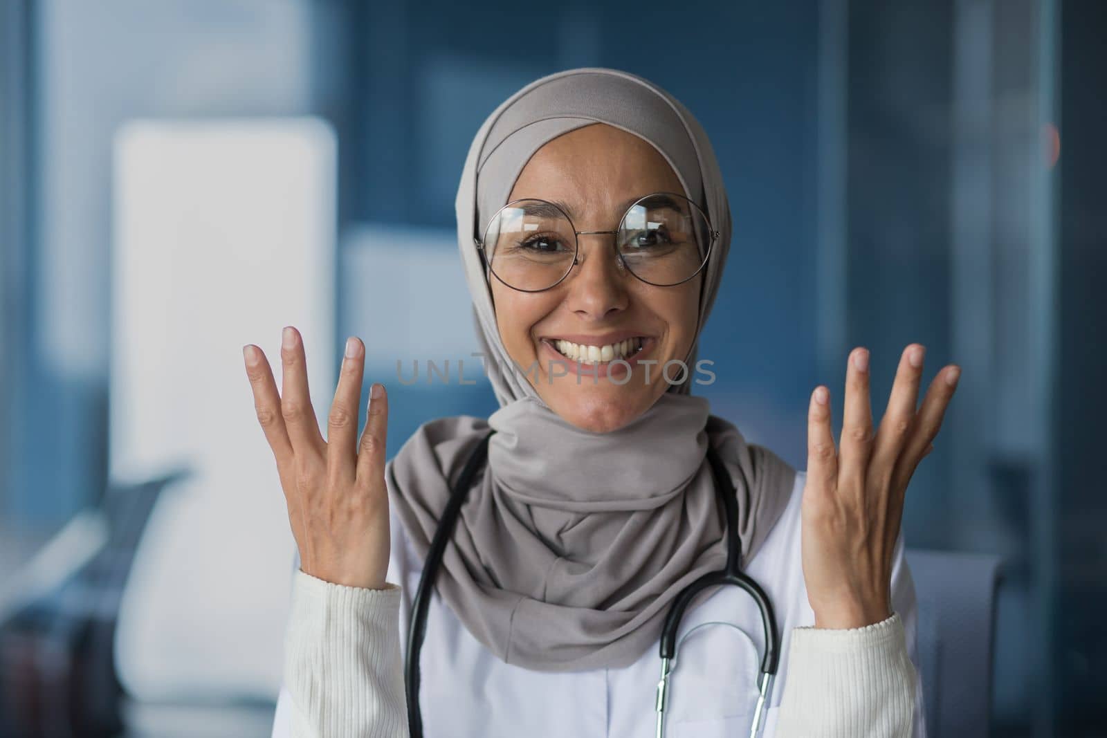 Happy and friendly young beautiful woman doctor student Muslim, Arabic. Standing in hijab, glasses and with stethoscope, looking at camera, pointing with hands.