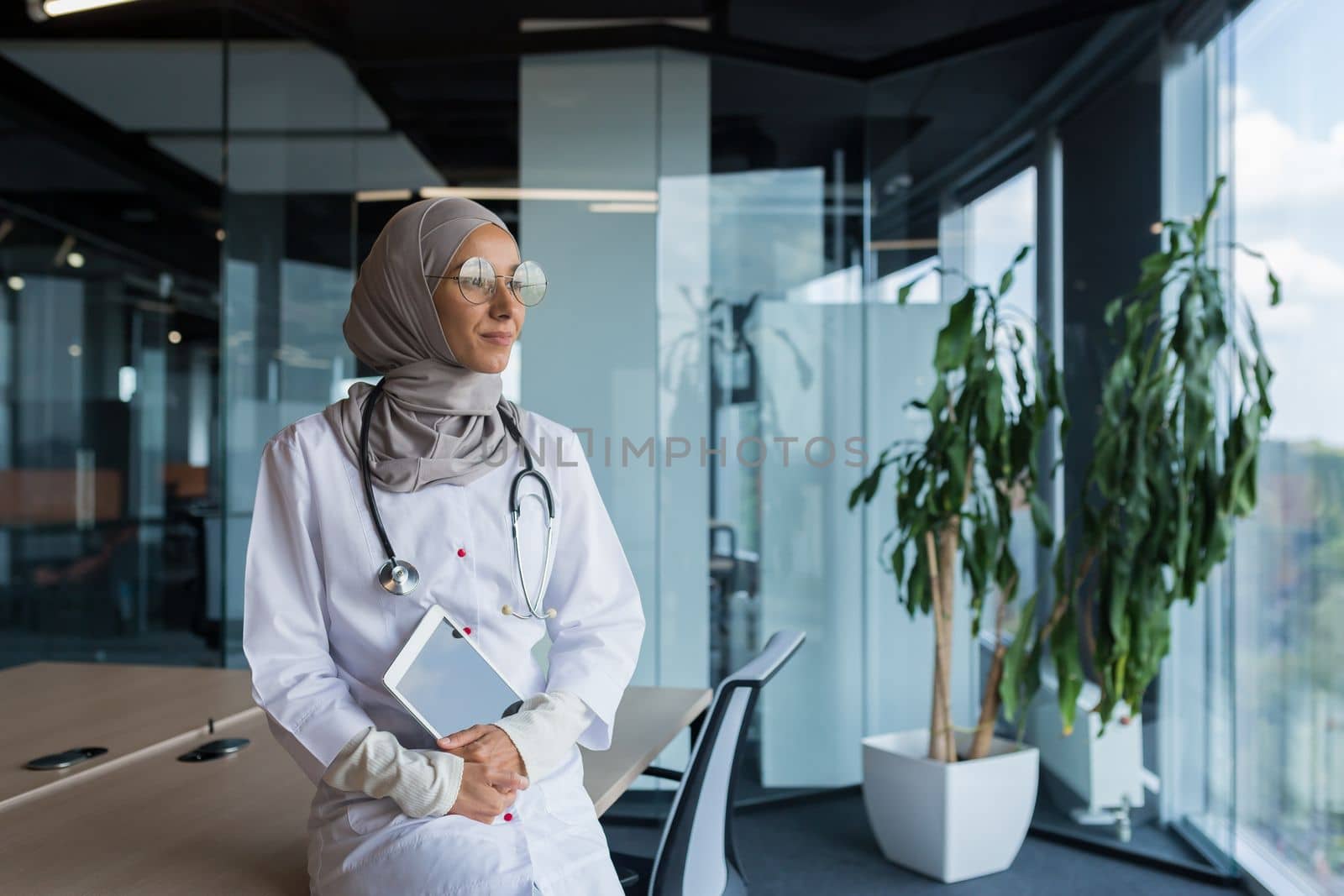 A happy and thoughtful young Muslim woman, an Arab doctor in a hospital, is sitting at her desk, holding a tablet in her hands, looking out the window, smiling, loving her work.