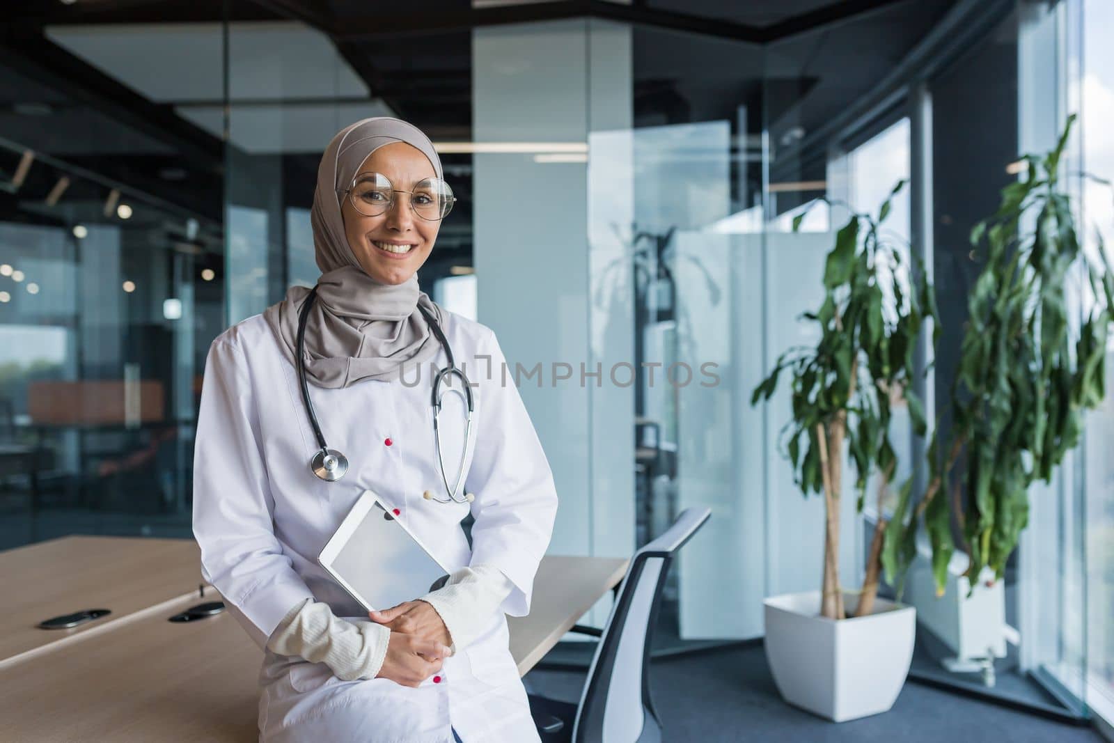 Portrait of a young happy and beautiful Muslim Arab female doctor, student sitting on a table in a large office in a hospital, holding a tablet in her hands, looking at the camera, smiling.