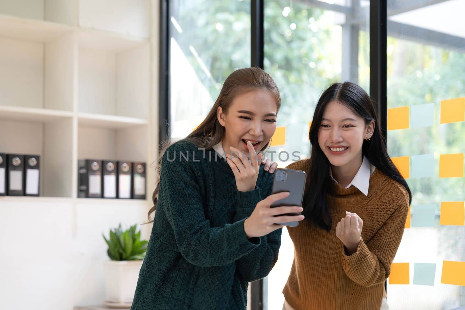 Two young Asian businesswomen show joyful expression of success at work smiling happily with a laptop computer in a modern office. by wichayada