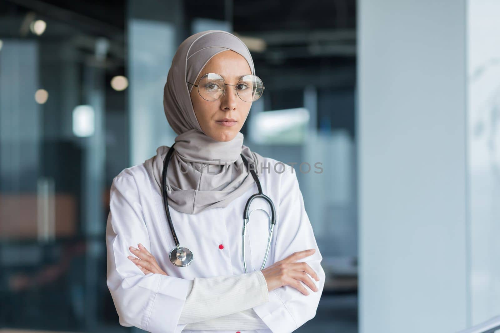Portrait of a young Muslim woman doctor. She is standing in the hall of the hospital dressed in a hijab, wearing glasses and a stethoscope, looking seriously into the camera, her arms crossed.
