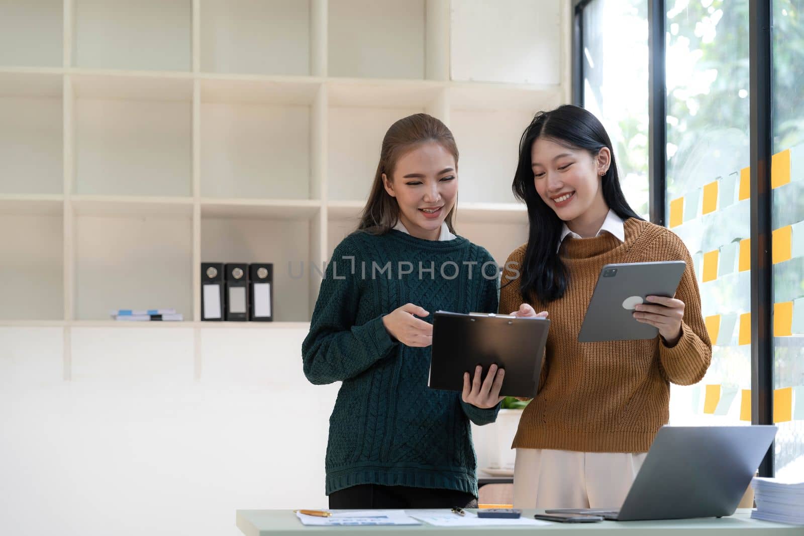 Two beautiful Asian businesswomen standing using digital tablet consulting and analyzing information in office work. by wichayada