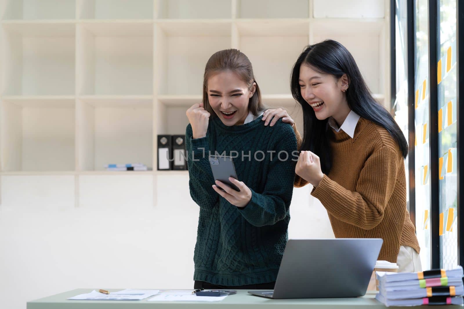 Two young Asian businesswomen show joyful expression of success at work smiling happily with a laptop computer in a modern office. by wichayada