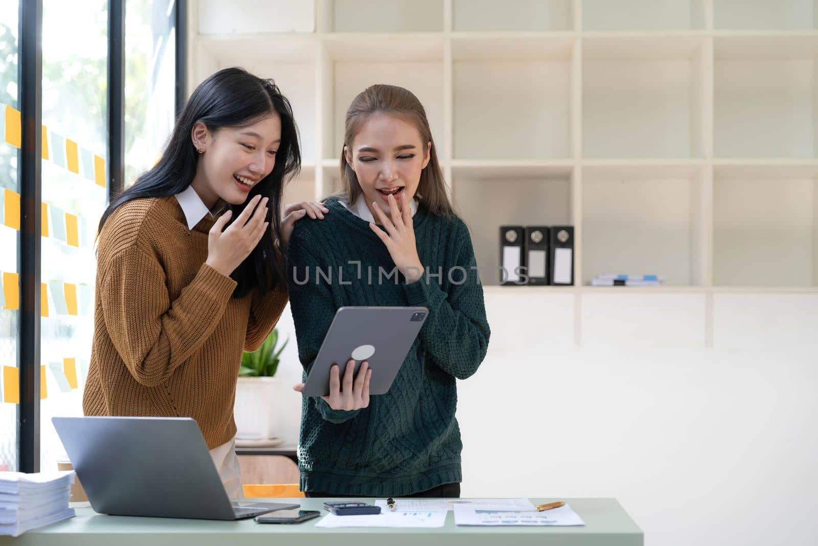 Two young Asian businesswomen show joyful expression of success at work smiling happily with a laptop computer in a modern office. by wichayada