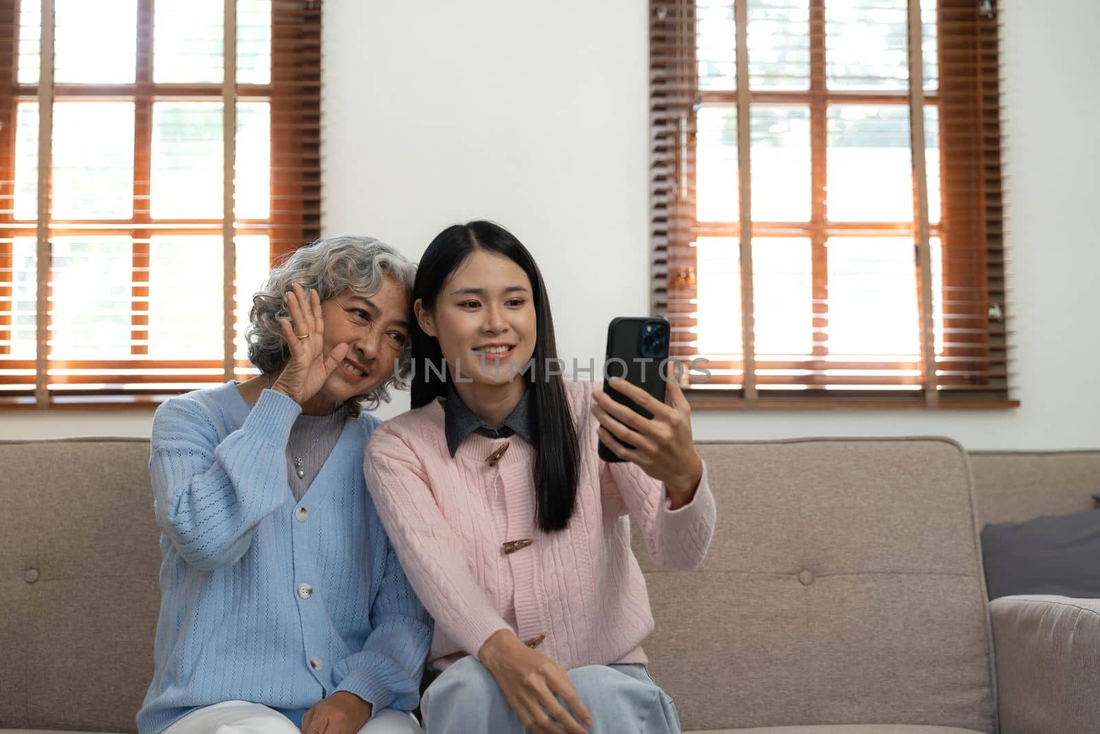 Smiling older mother and adult daughter using phone together, sitting on cozy sofa at home, happy young woman and mature mum looking at smartphone screen, watching video, having fun, two generations..