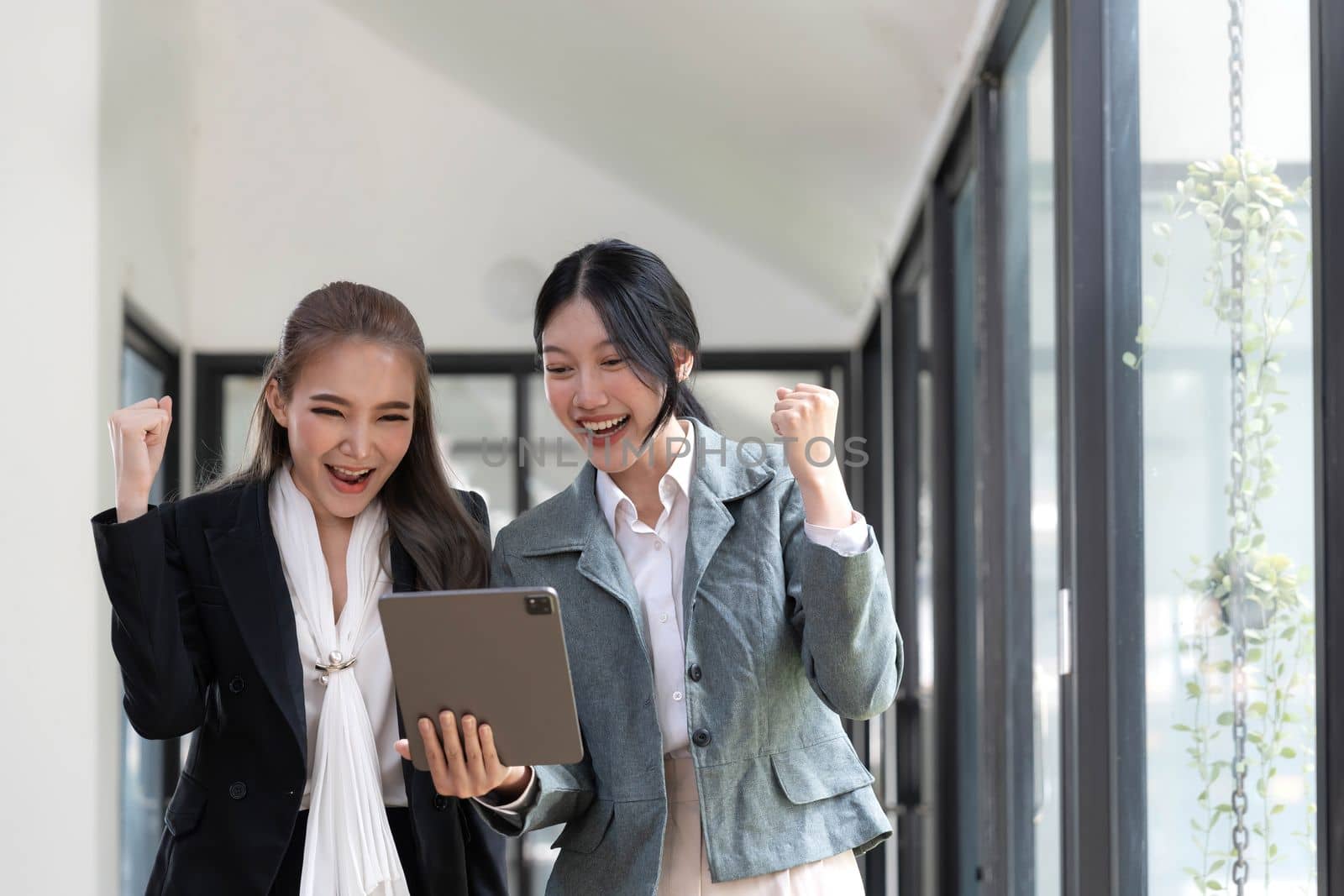 Two beautiful Asian businesswomen standing using digital tablet consulting and analyzing information in office work. by wichayada