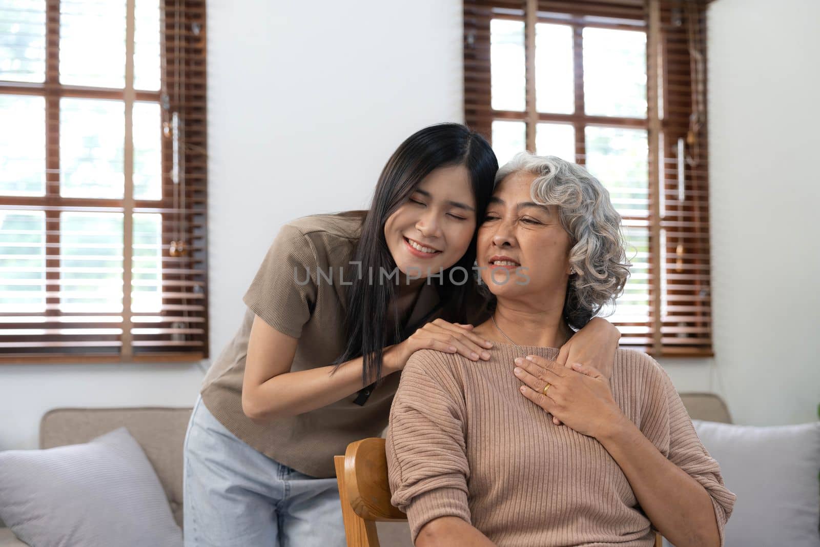 Loving adult daughter hugging older mother, standing behind couch at home, family enjoying tender moment together, young woman and mature mum or grandmother looking at each other, two generations..