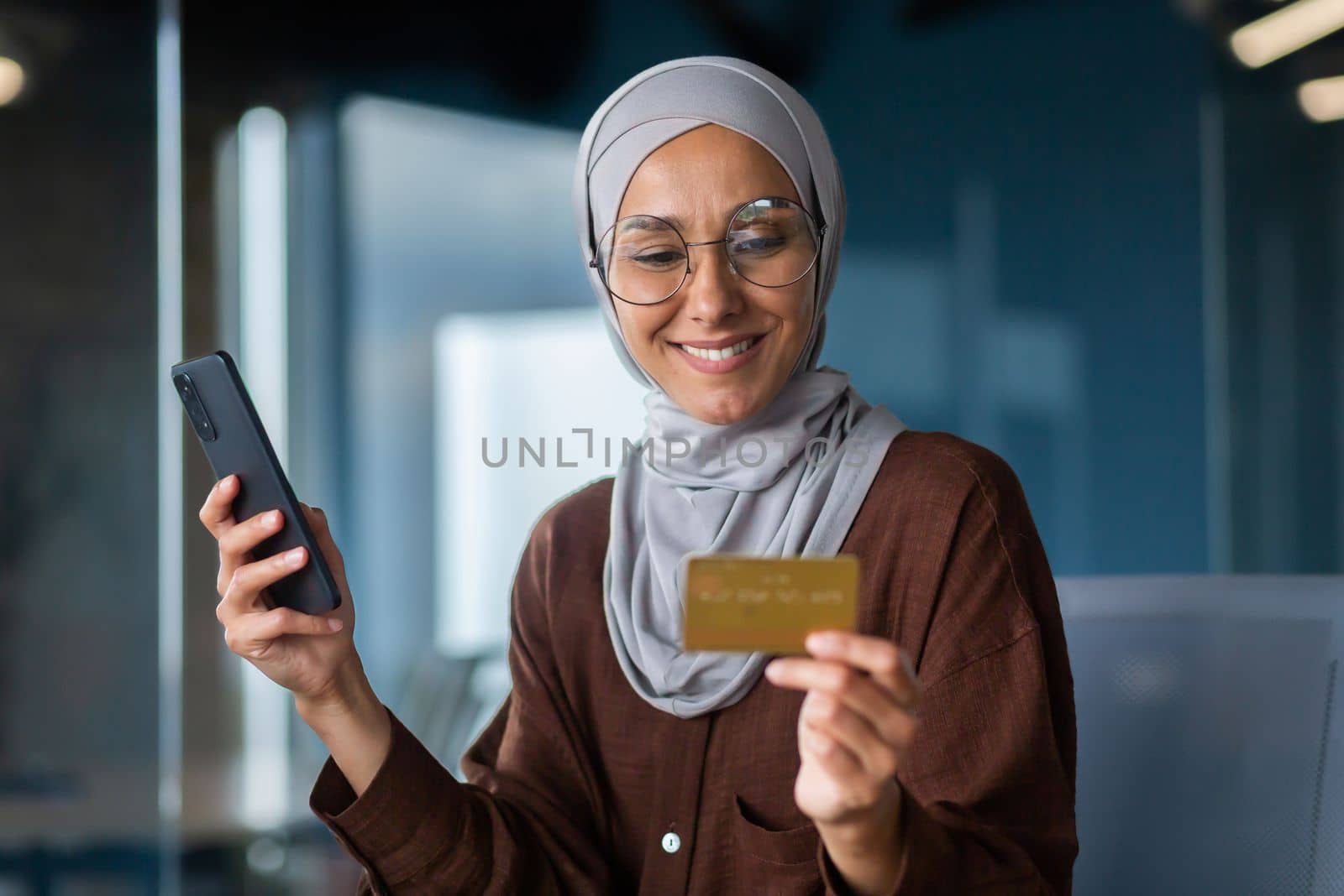 Successful businesswoman in hijab working inside office with laptop at workplace, muslim woman holding phone and bank credit card for online shopping and money transfer.
