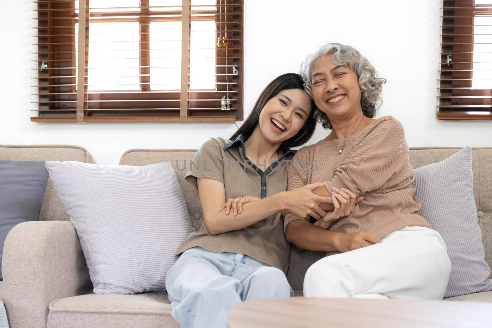 Portrait of Asian lovely family, young daughter hugging older mother. Attractive woman and senior elder mature mom sit on sofa, enjoy activity at home together in living room and smile, look at camera by wichayada