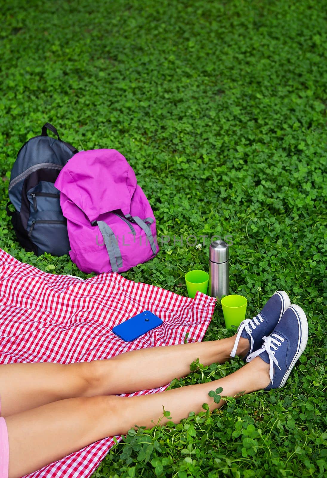 Top view of a young female student sitting on a plaid blanket and green grass in the park. Break in the open air, drink coffee, tea, phone in hand