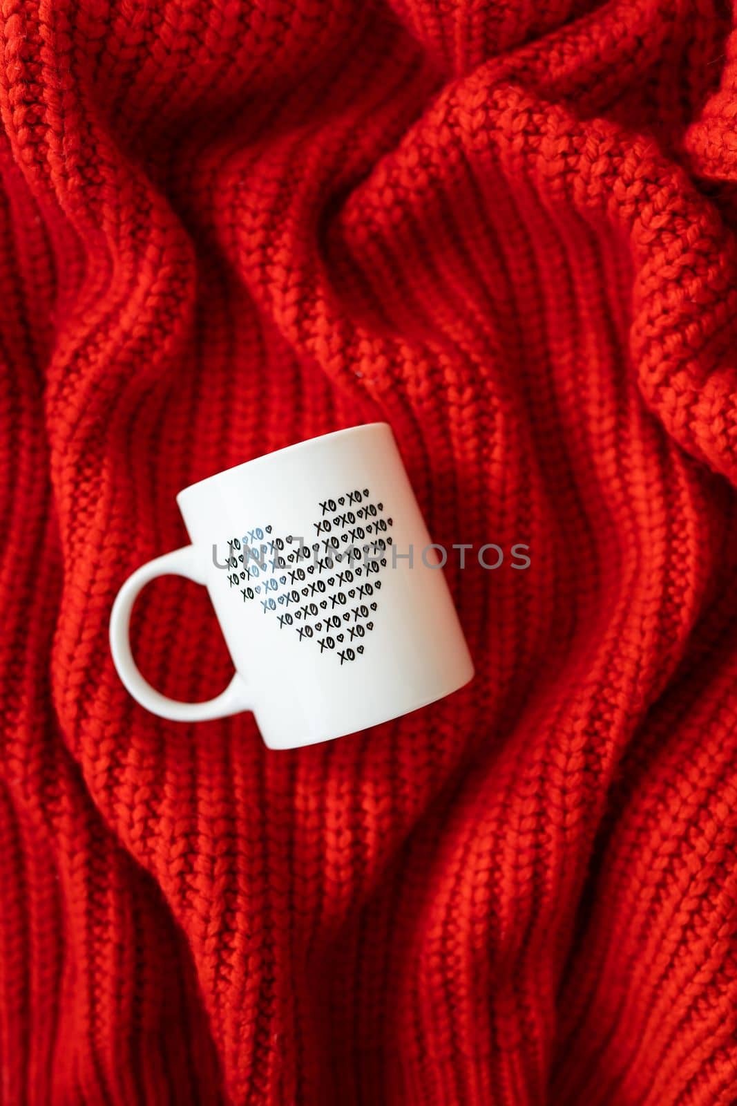 A white cup with a heart stands on a red knitted fabric. The concept of love and celebration of St. Valentine's day, February 14th