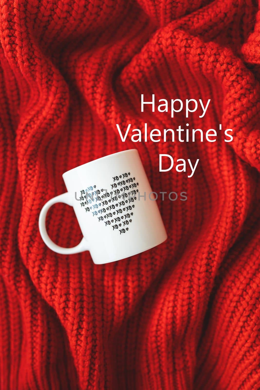 A white cup with a heart stands on a red knitted fabric. Inscription of St. Valentine's day, 14 February