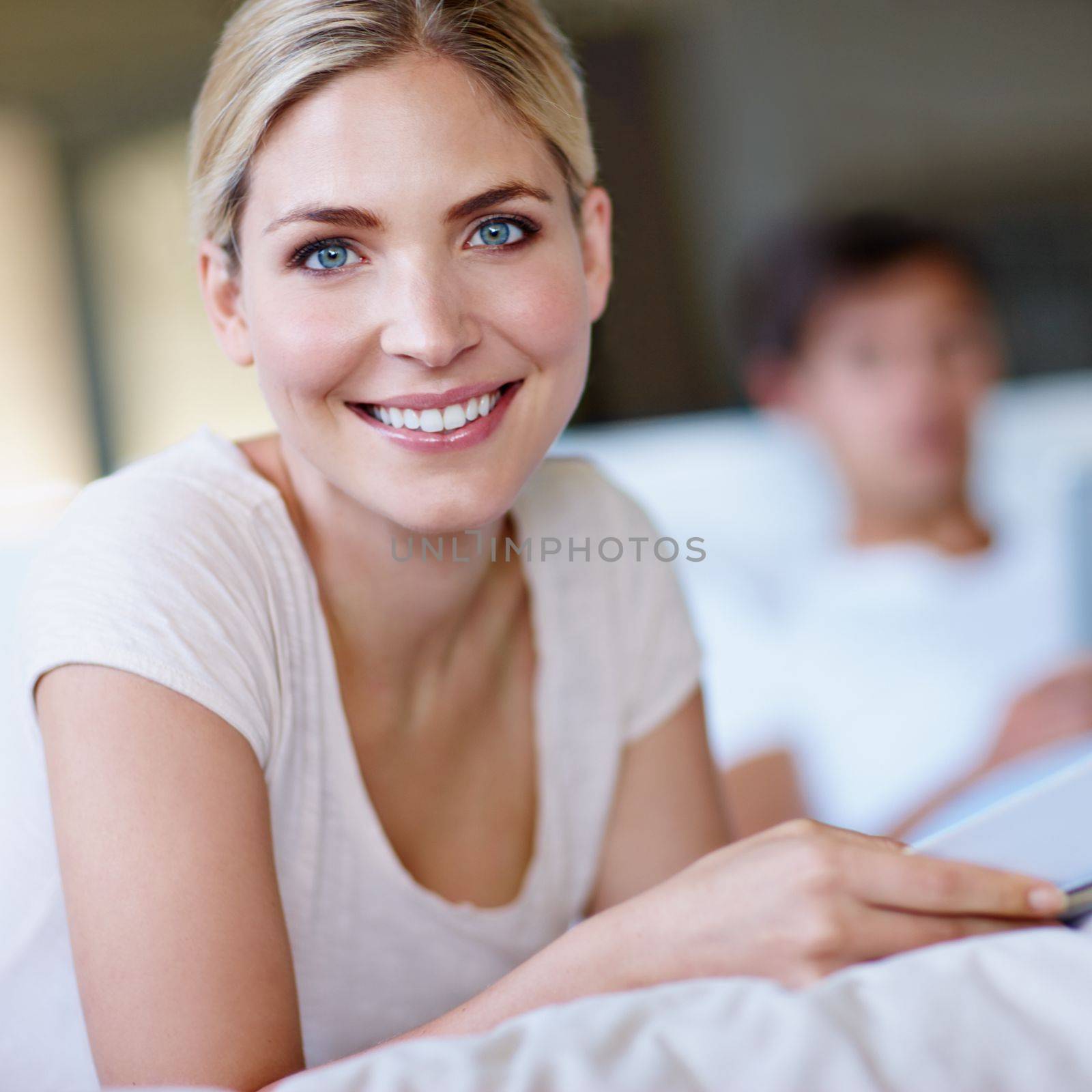 Happiness is an inside job. a happy young woman lying in bed and reading a magazine with her husband in the background. by YuriArcurs