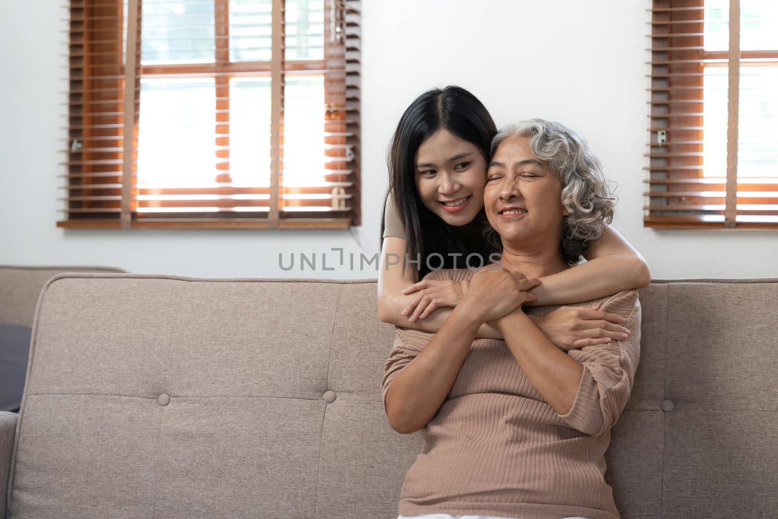 Loving adult daughter hugging older mother, standing behind couch at home, family enjoying tender moment together, young woman and mature mum or grandmother looking at each other, two generations by wichayada