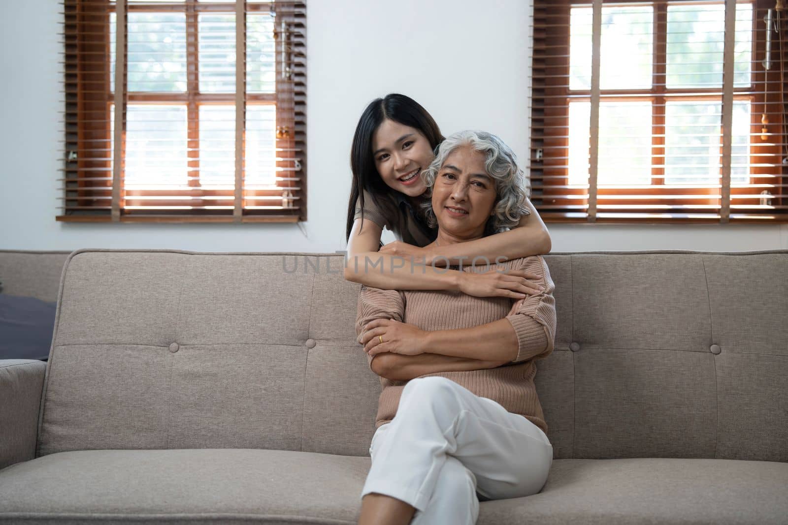 Loving adult daughter hugging older mother, standing behind couch at home, family enjoying tender moment together, young woman and mature mum or grandmother looking at each other, two generations by wichayada