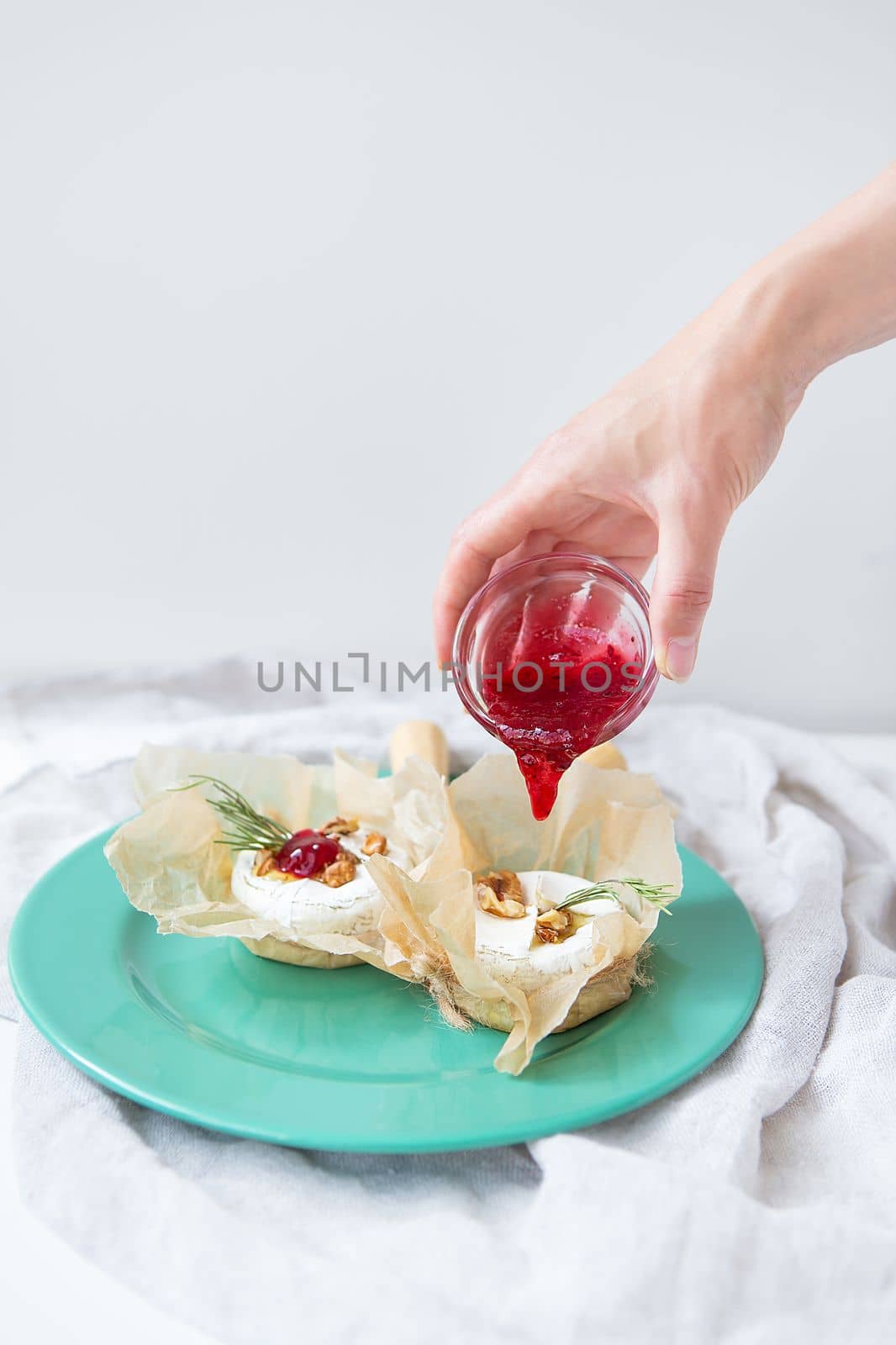 Baked camembert with walnuts, honey and rosemary wrapped in parchment lies on a green plate, a girl pours cranberry jam into the cheese, delicious and healthy food. by sfinks